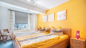 A bedroom with a bed, bedside table, chair, and window with curtains. - Apartment 'Fewo Bauer Mecke' with Shared Garden (Mirow)