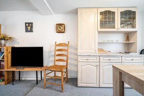 Een kamer met een houten tv-meubel, een rieten stoel, een keuken met witte kasten en een eettafel.