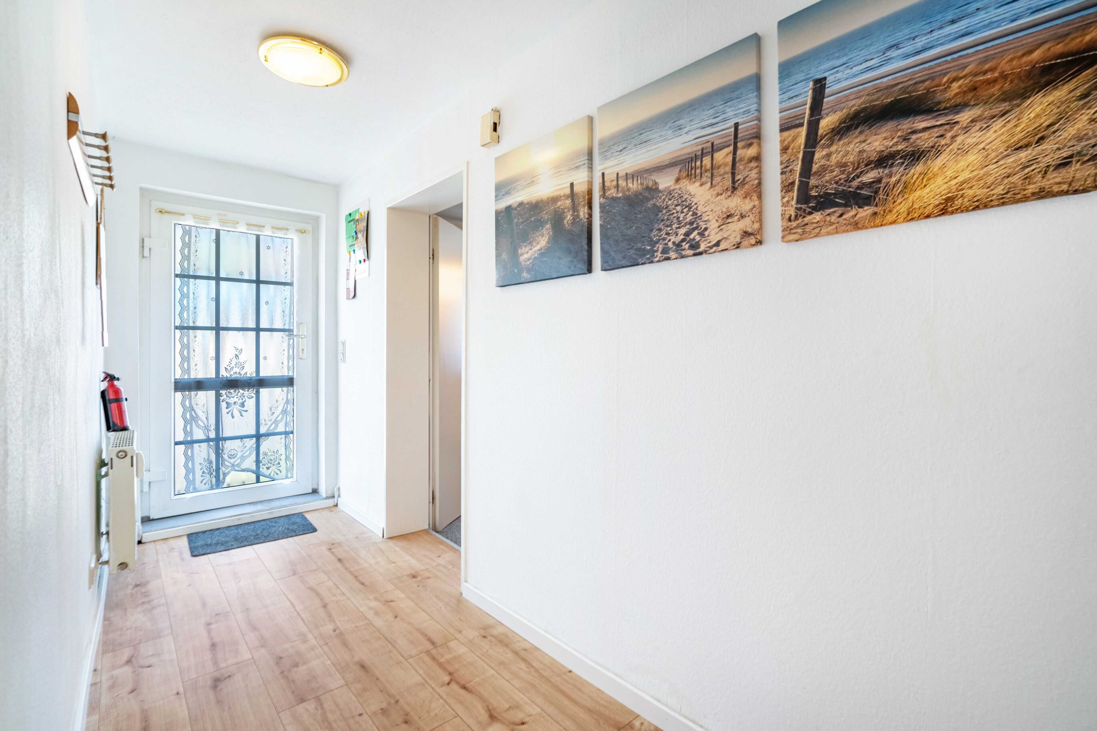 A hallway with wooden flooring, a door with a decorative glass panel, and three wall-mounted pictures depicting coastal scenes.