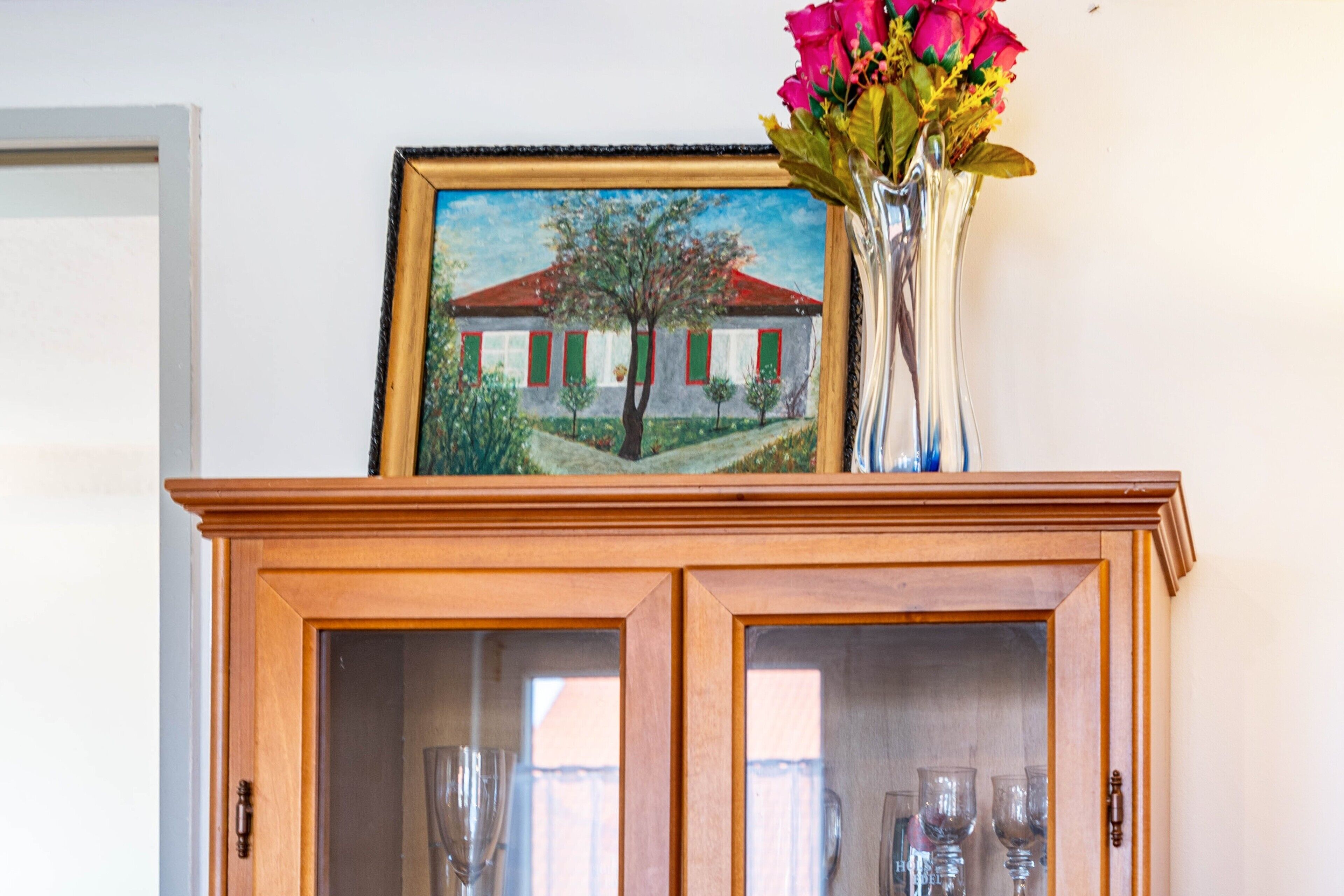 A wooden cabinet with glass doors, a framed painting of a house, and a vase with flowers on top.