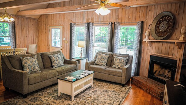 A cozy living room with wooden walls, two sofas with patterned cushions, a coffee table, a fireplace, and a ceiling fan. - Recently renovated, beautiful chalet just miles from Wild Animal Safari (Pine Mountain)
