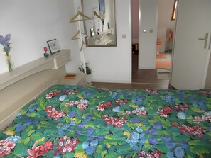 Un dormitorio con una colcha estampada, un espejo, un armario y una puerta que da a otra habitación.