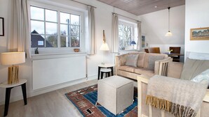 Una sala de estar con sofá, reposapiés y lámpara, ventanas amplias y vistas al exterior.