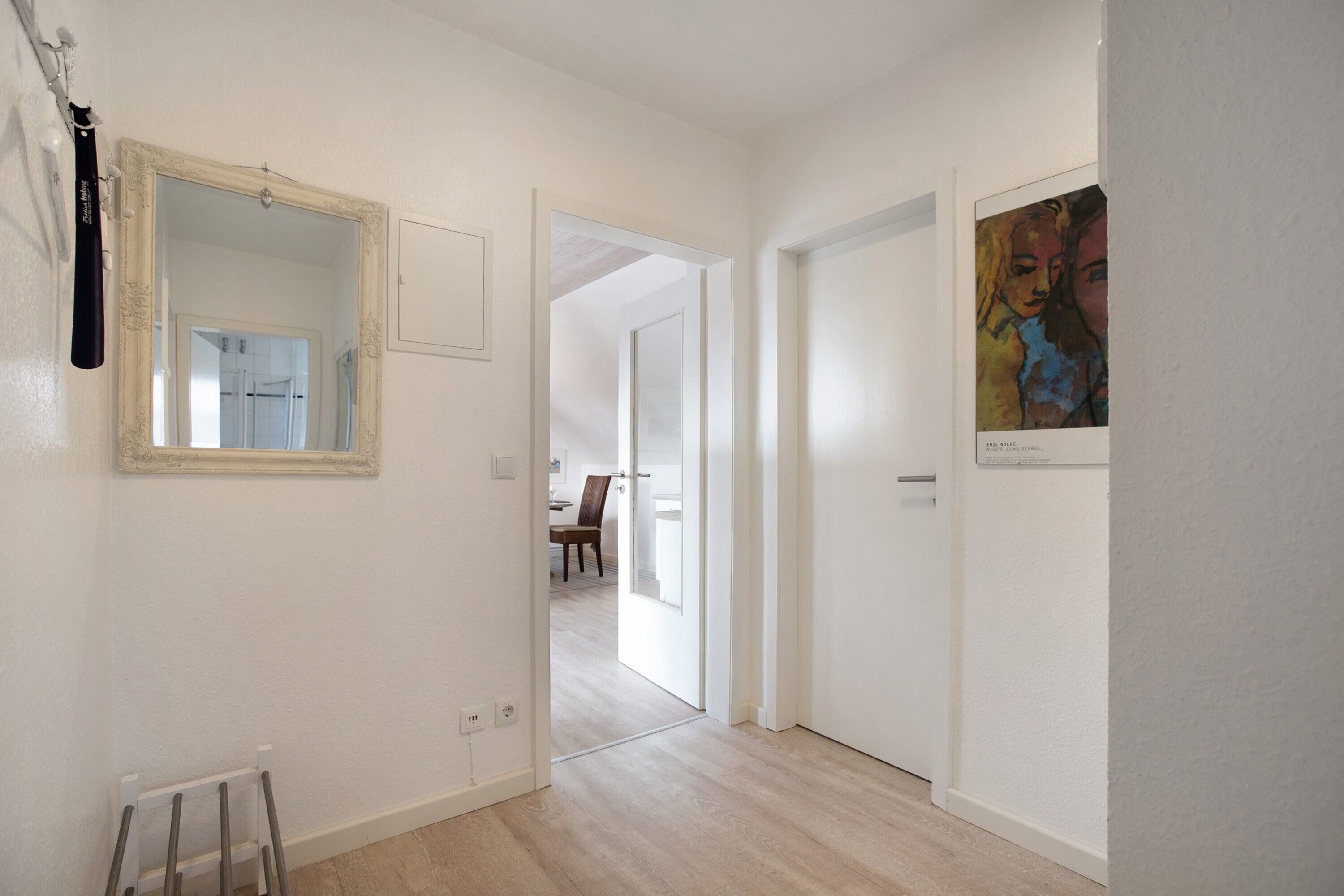 Un couloir avec un miroir, une œuvre d’art encadrée et une porte donnant sur une autre pièce.