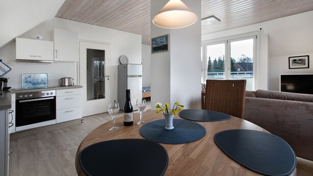 Una cocina moderna con armarios blancos, horno empotrado y zona de comedor con mesa de madera y posavasos negros.