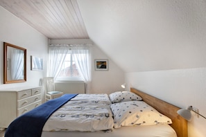 A bedroom with a bed, a chair, a dresser, a window with curtains, and a wall-mounted lamp. - Apartment 'Ferienwohnung Ackerkoog' with Balcony and Wi-Fi (Niebüll)