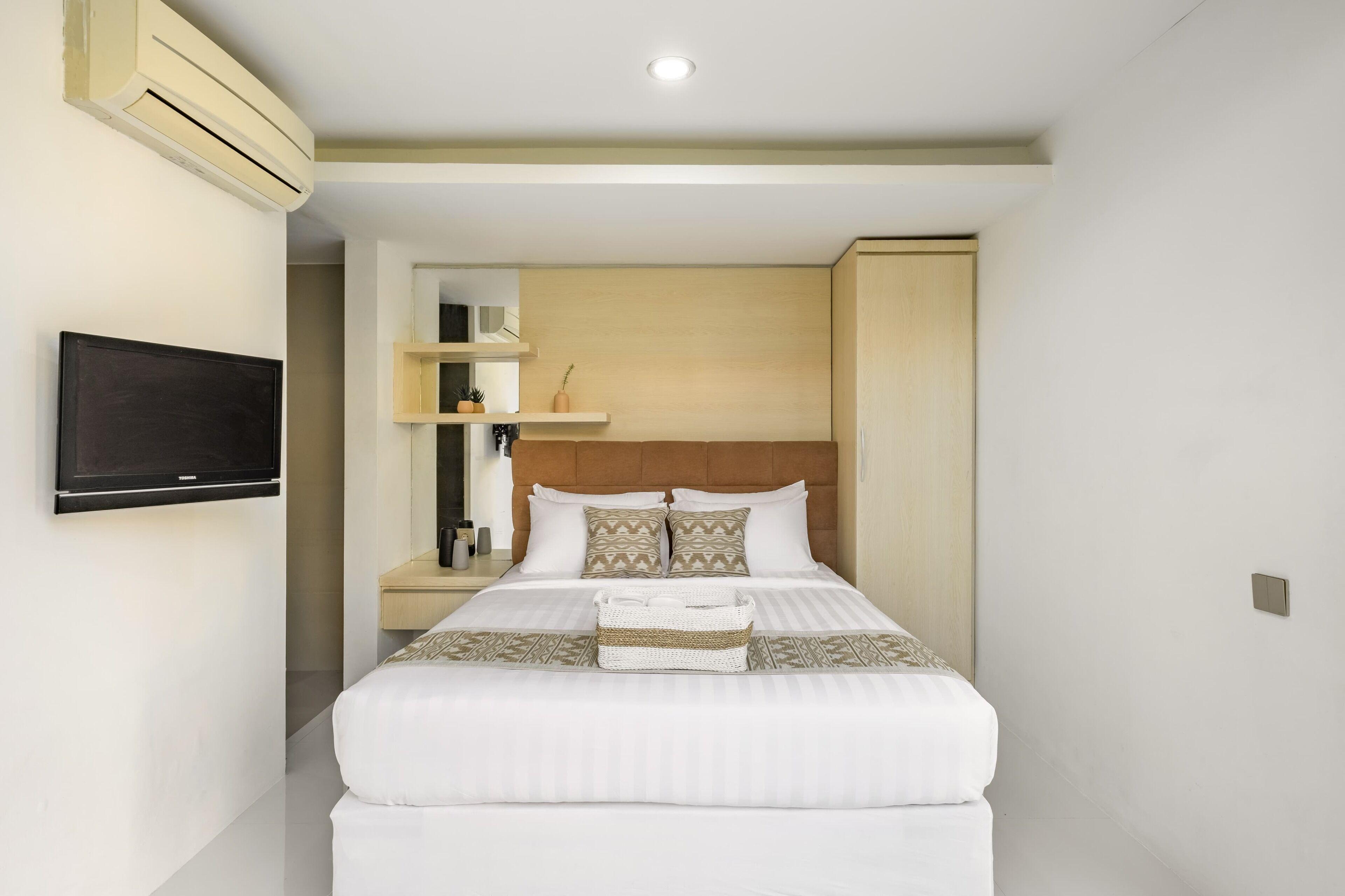 A hotel room with a bed, a television mounted on the wall, a wooden headboard, and a shelf with decorative items.