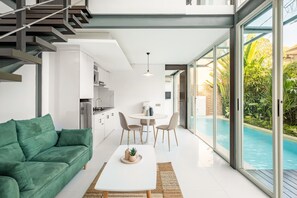 A modern living room with a green sofa, a white coffee table, and a dining area with a table and chairs. A large window reveals a swimming pool and lush greenery outside. - Villa De Luca By Aishwarya Bali International (Kerobokan)