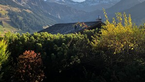 Una cabaña de montaña con techo inclinado, rodeada de un bosque denso y montañas nevadas al fondo.