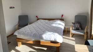 Un dormitorio con una cama de madera, dos sillas grises, una mesita redonda y un estante fijado a la pared.