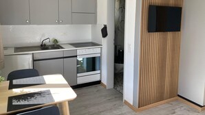 Una cocina moderna con gabinetes grises, un horno integrado y un fregadero. Hay un comedor con mesa y sillas, y un televisor montado en la pared.