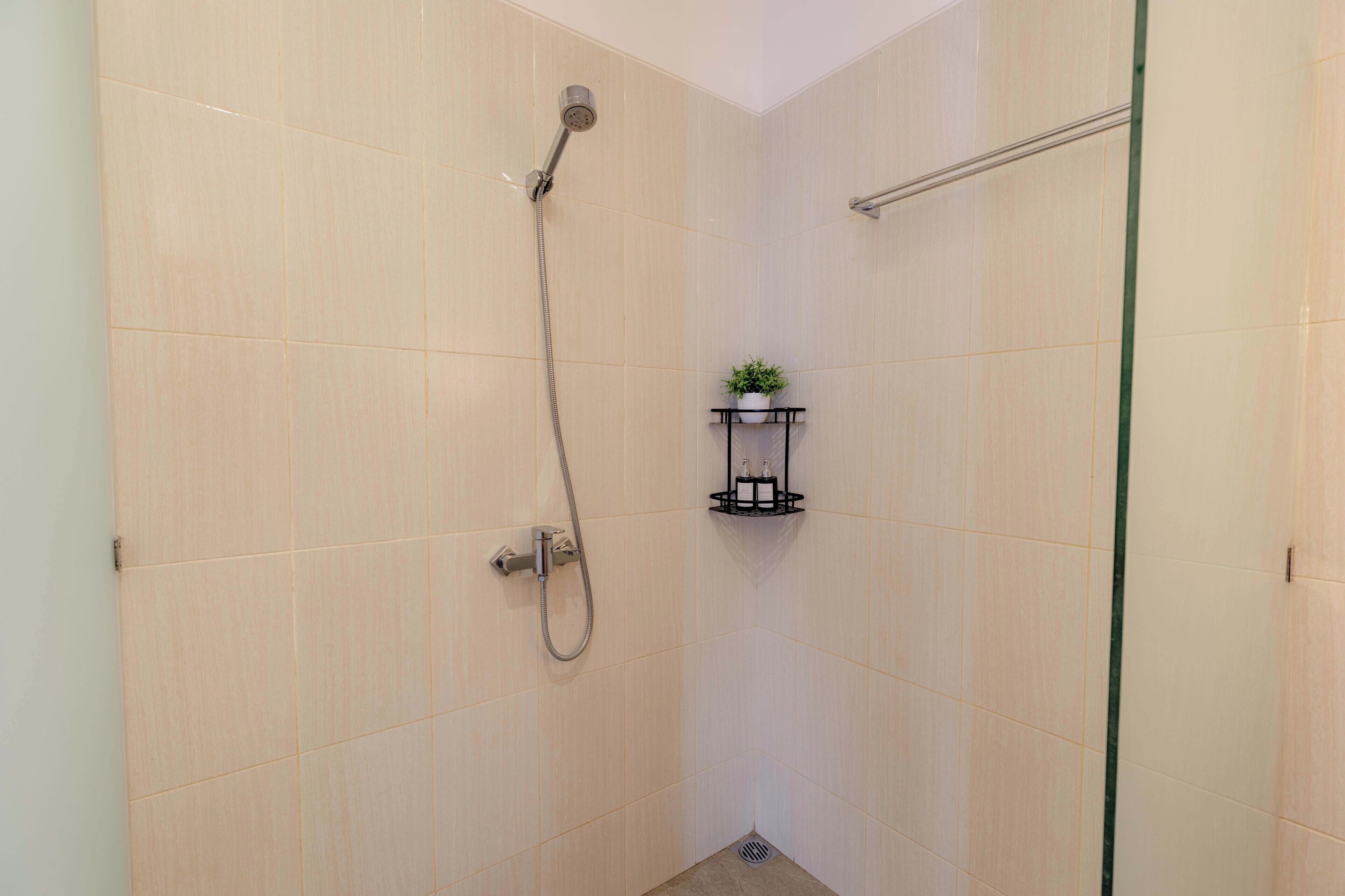 Ein kleiner Duschbereich mit gefliestem Boden, einer Handbrause und einem an der Wand montierten Wasserhahn. Es gibt ein kleines Regal mit einer Pflanze und einem Seifenspender.