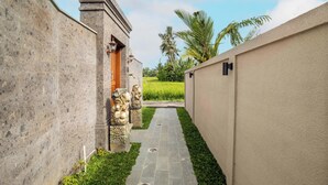 A narrow pathway flanked by stone walls and a grassy area with palm trees in the background. - Weda Villas (Ubud)