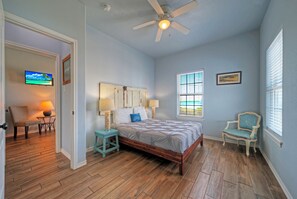 A bedroom with a wooden floor, a bed with a headboard, a ceiling fan, a TV, and two chairs.