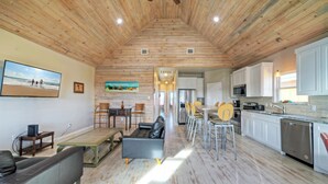 A spacious living area with a wooden ceiling, a dining table with chairs, a sofa, and a kitchen with white cabinets and appliances. - Sleeps 46, 6 Homes | Private Boat Dock, Waterfront Pool, Views | Laguna Village (Corpus Christi)