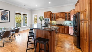 A spacious kitchen with wooden cabinets, a central island with stools, and a dining area with chairs. - Sunset Pointe by AvantStay | Waterfront, Beach, Fire Pit | Sleeps 14 | 5BDR 4BA (Troutman)