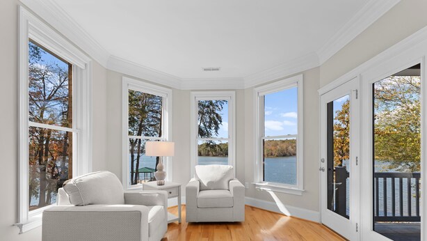 A spacious room with hardwood flooring, a white sofa, a chair, and a table with a lamp. Large windows offer views of trees and a body of water. - Sunset Pointe by AvantStay | Waterfront, Beach, Fire Pit | Sleeps 14 | 5BDR 4BA (Troutman)