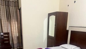 A neatly made bed with a white mattress and a purple blanket, a wooden wardrobe, a chair, and a curtain with a geometric pattern. - Hotel Blessing Inn (Jalandhar)