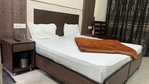 A neatly made bed with white pillows and a brown blanket, a wooden nightstand with a remote control, and a wall socket. - Hotel Blessing Inn (Jalandhar)