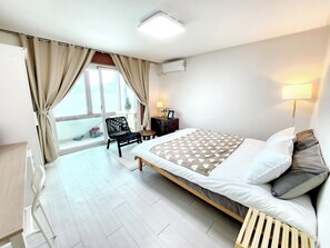 Quarto de hotel com cama, poltrona, escrivaninha, luminária e janela com cortinas.