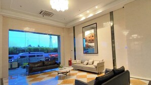 A modern lobby with a checkered floor, a large window with a view of parked cars, a sofa, a coffee table, and a framed picture on the wall. - Sky House Bsd By Mizu Rooms (Pagedangan)