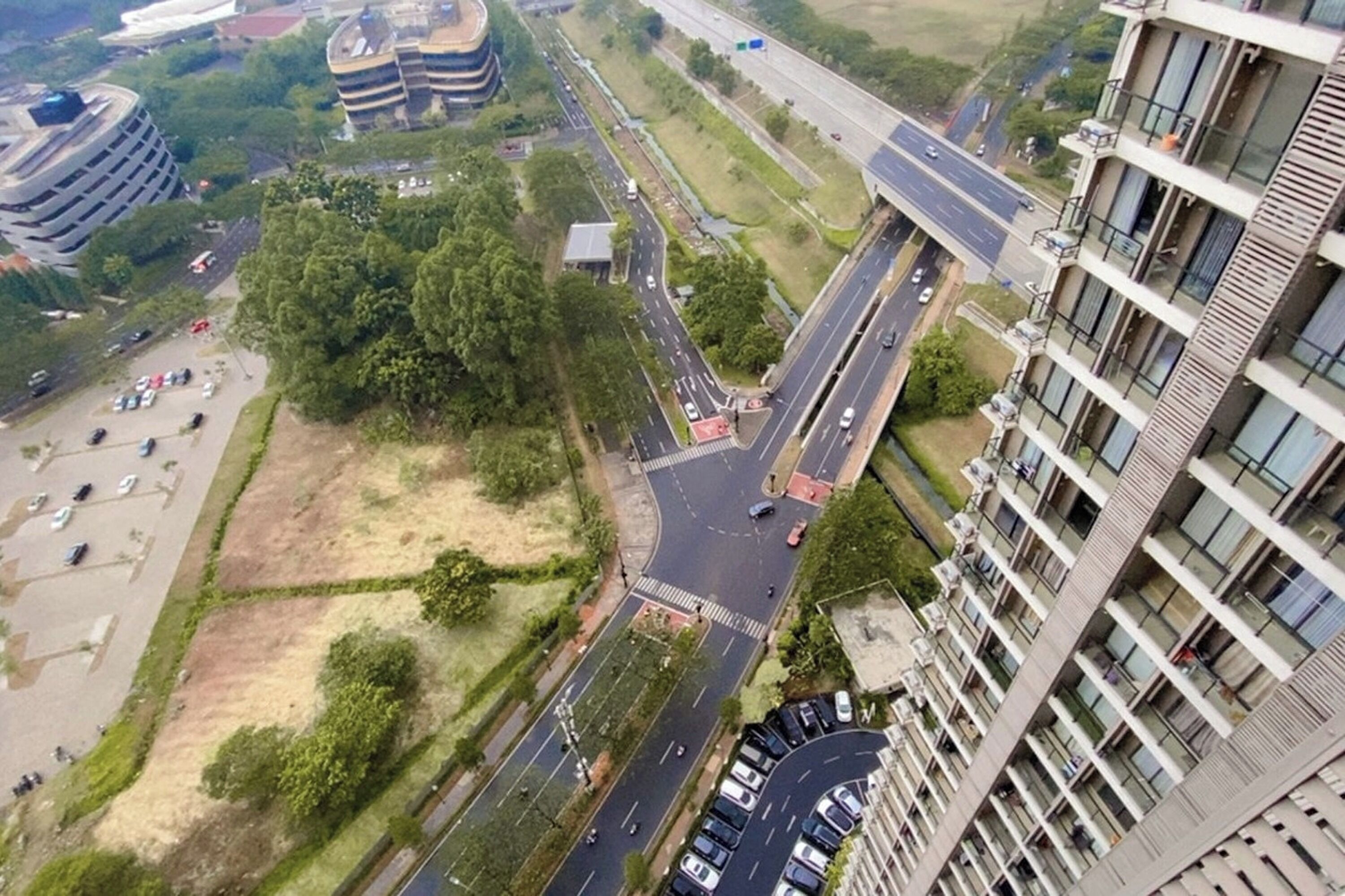 Une vue aérienne d’un parking à plusieurs niveaux situé à côté d’une autoroute, avec de la végétation environnante et un grand bâtiment en arrière-plan.