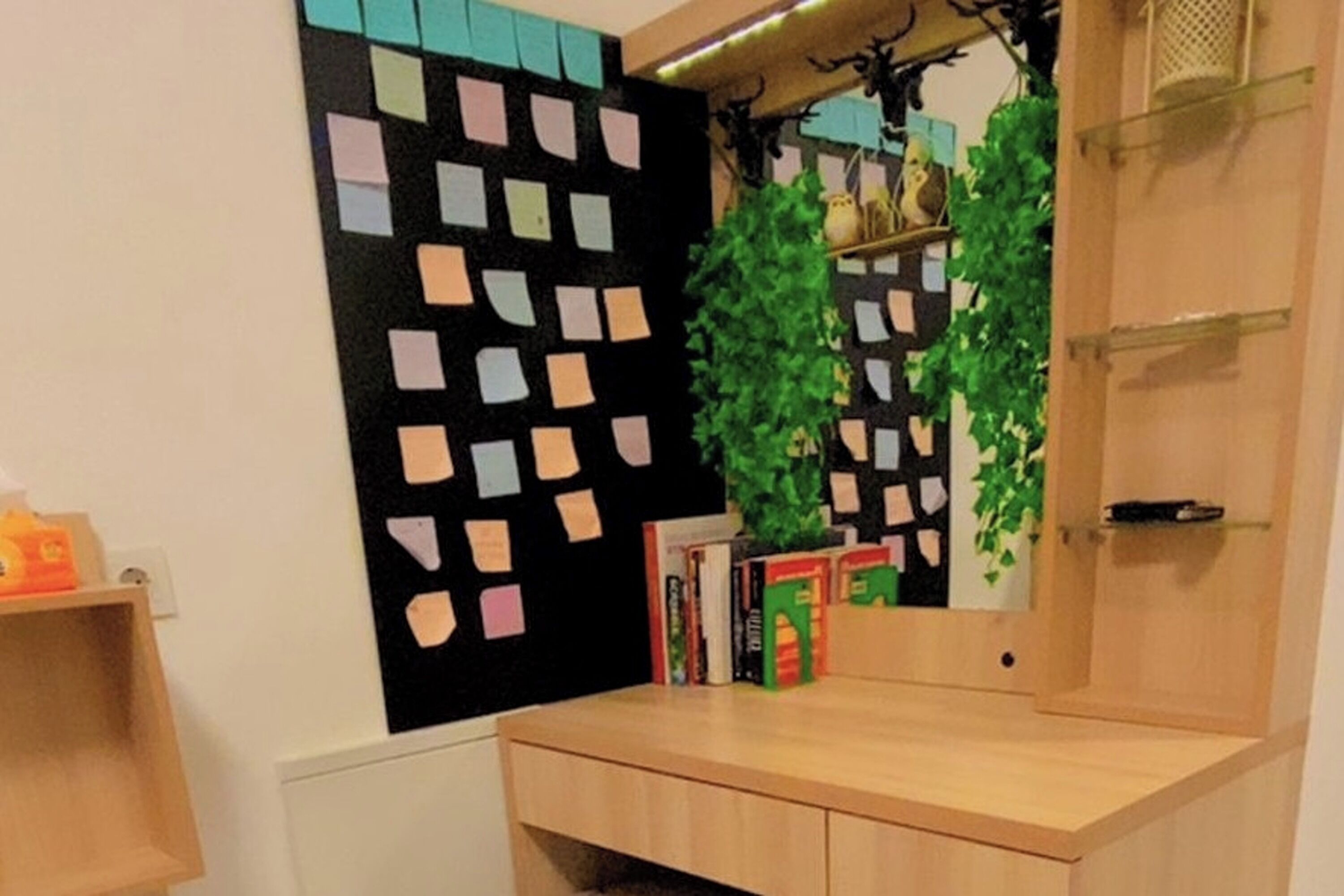 A room with a wooden desk, a blackboard with colorful sticky notes, and a mirror. There are books and a small shelf with decorative items.