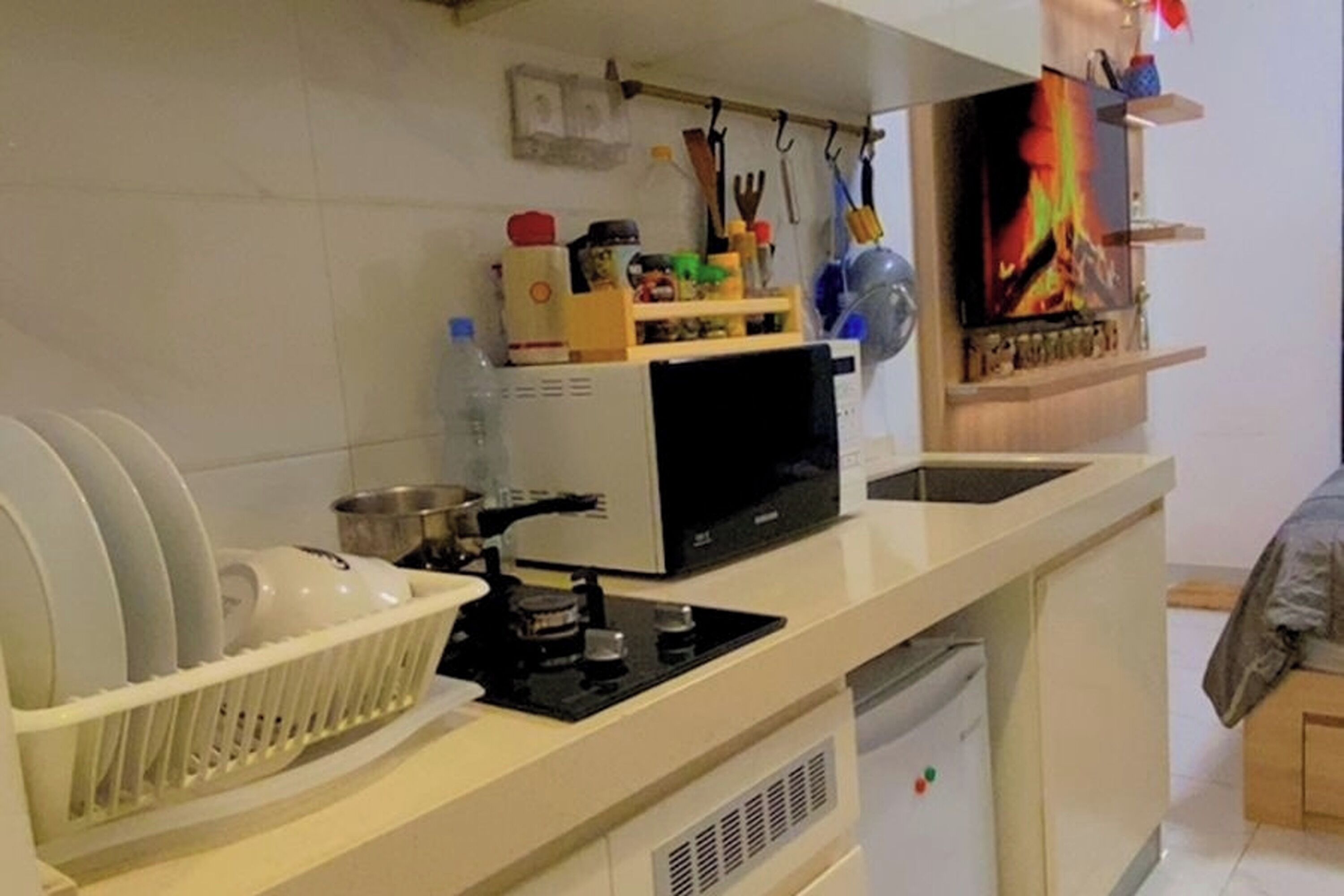 A compact kitchen with a microwave, sink, and dish rack. There is a small refrigerator and a shelf with various kitchen items.
