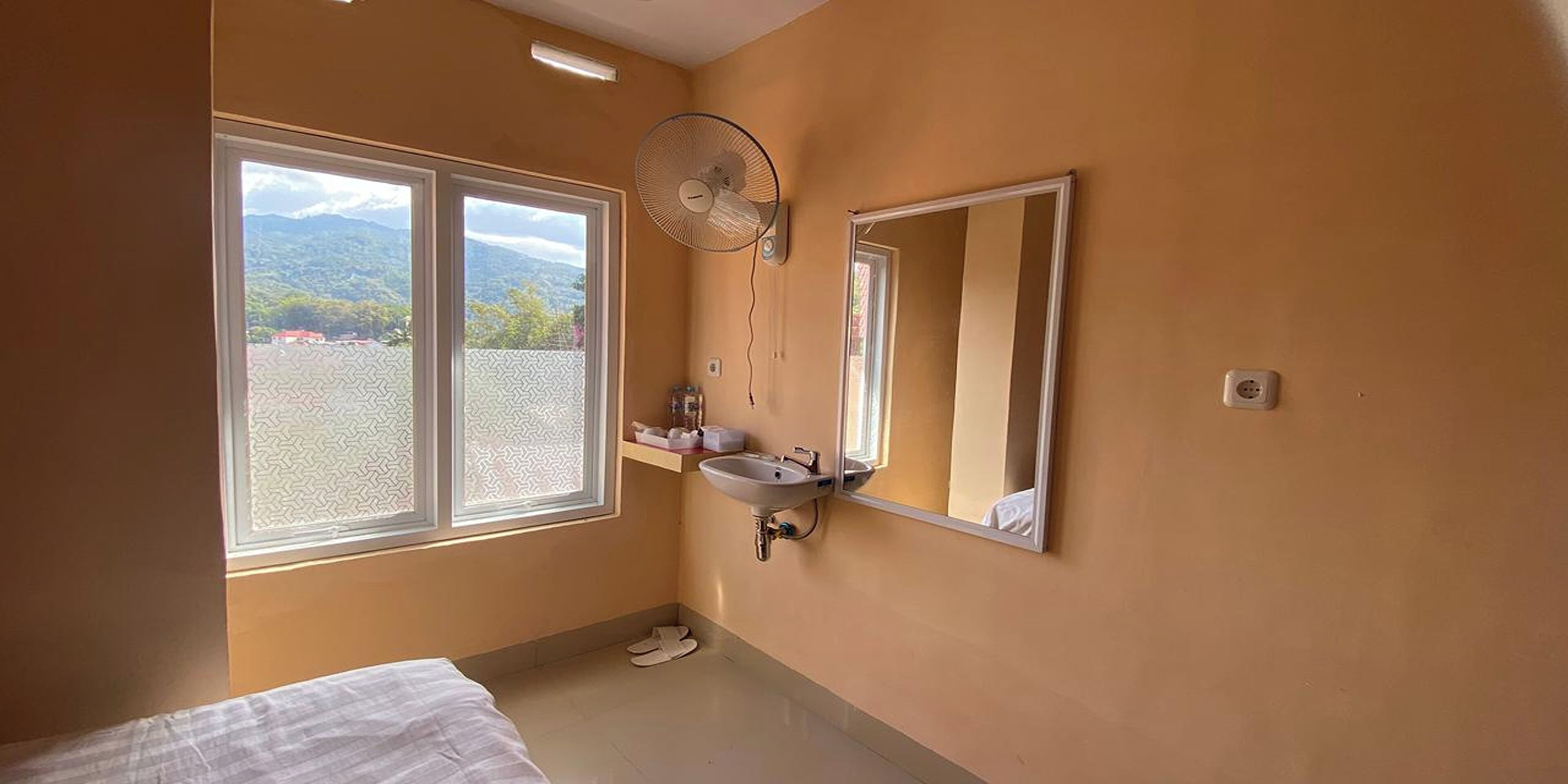 Quarto com cama, janela com vista para montanhas, ventilador e pia com espelho.