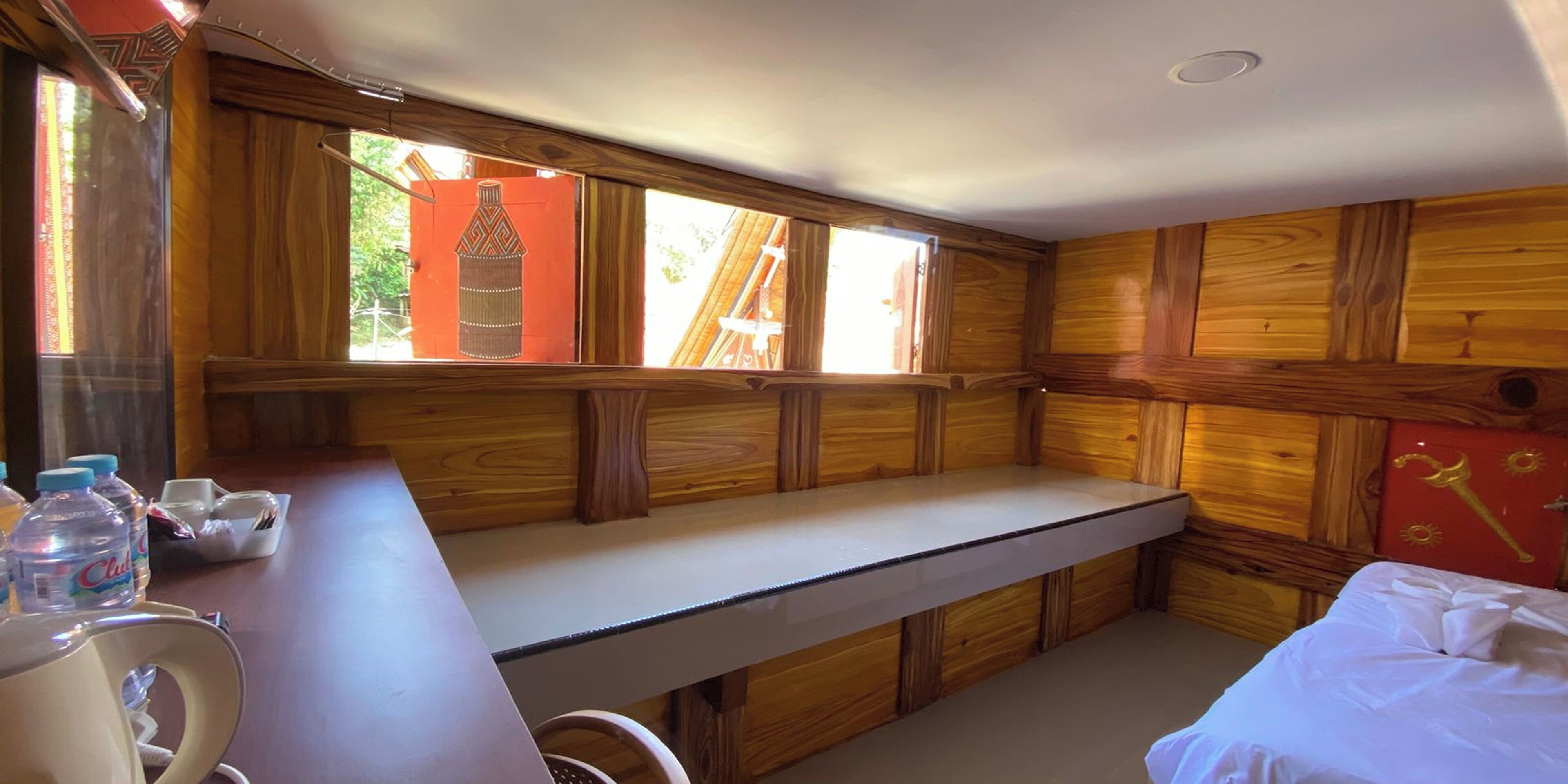 A room with wooden paneling, a white desk with a chair, a bed with white bedding, and a window with a view of a red structure outside.