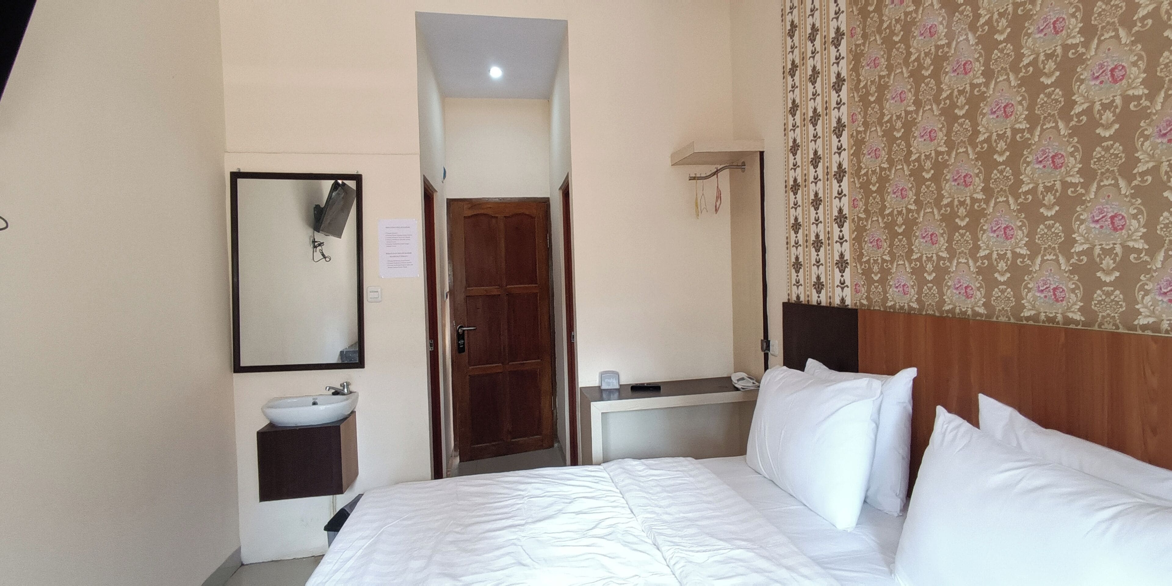 A hotel room with a bed, a bedside table, a mirror, a sink, and a door.