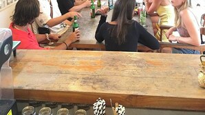 A group of people seated around a wooden bar with hanging wicker lamps, a patterned wall, and potted plants in the background. - Coco & Pineapple Pants Hostel (Canggu)