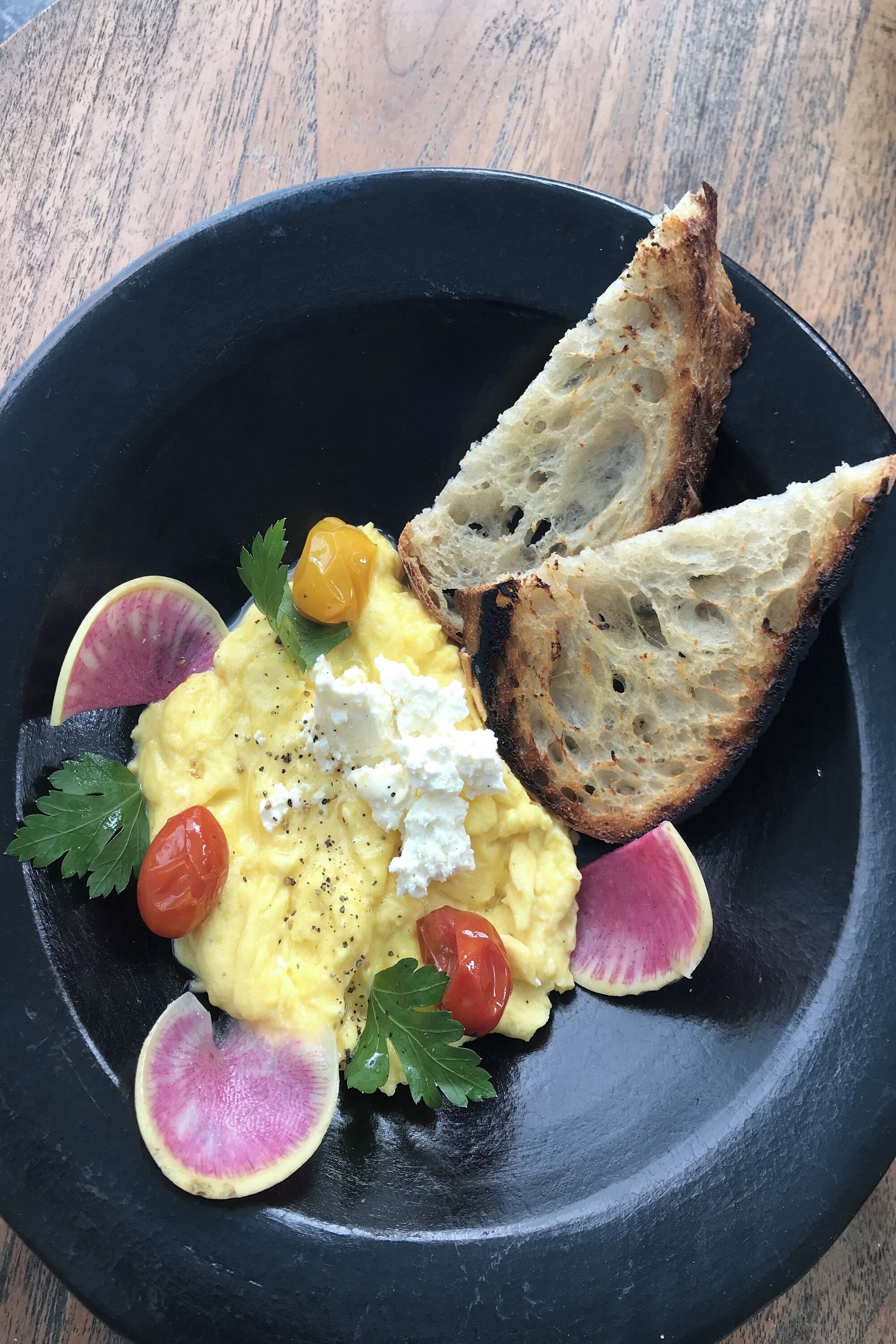 A black plate with scrambled eggs, feta cheese, cherry tomatoes, and toasted bread.