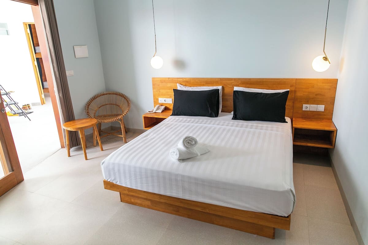 Un lit bien fait, avec une serviette enroulée, deux tables de chevet, une tête de lit en bois et une chaise en osier.