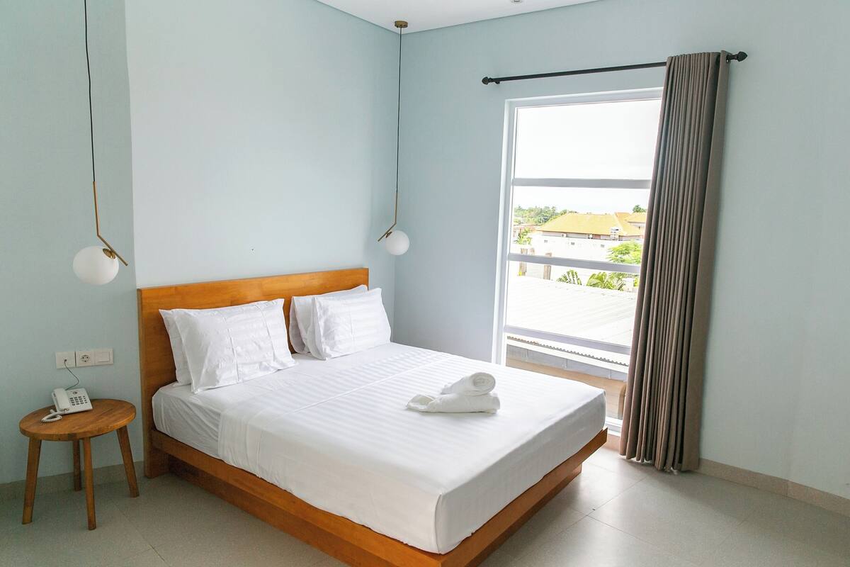 Un lit bien fait, avec du linge de lit blanc, une tête de lit en bois et une serviette blanche posée sur le lit. On y trouve une petite table en bois avec un téléphone et une lampe. Les murs de la pièce sont d’un bleu clair, il y a une grande fenêtre donnant sur les bâtiments à l’extérieur, et un rideau à la fenêtre.