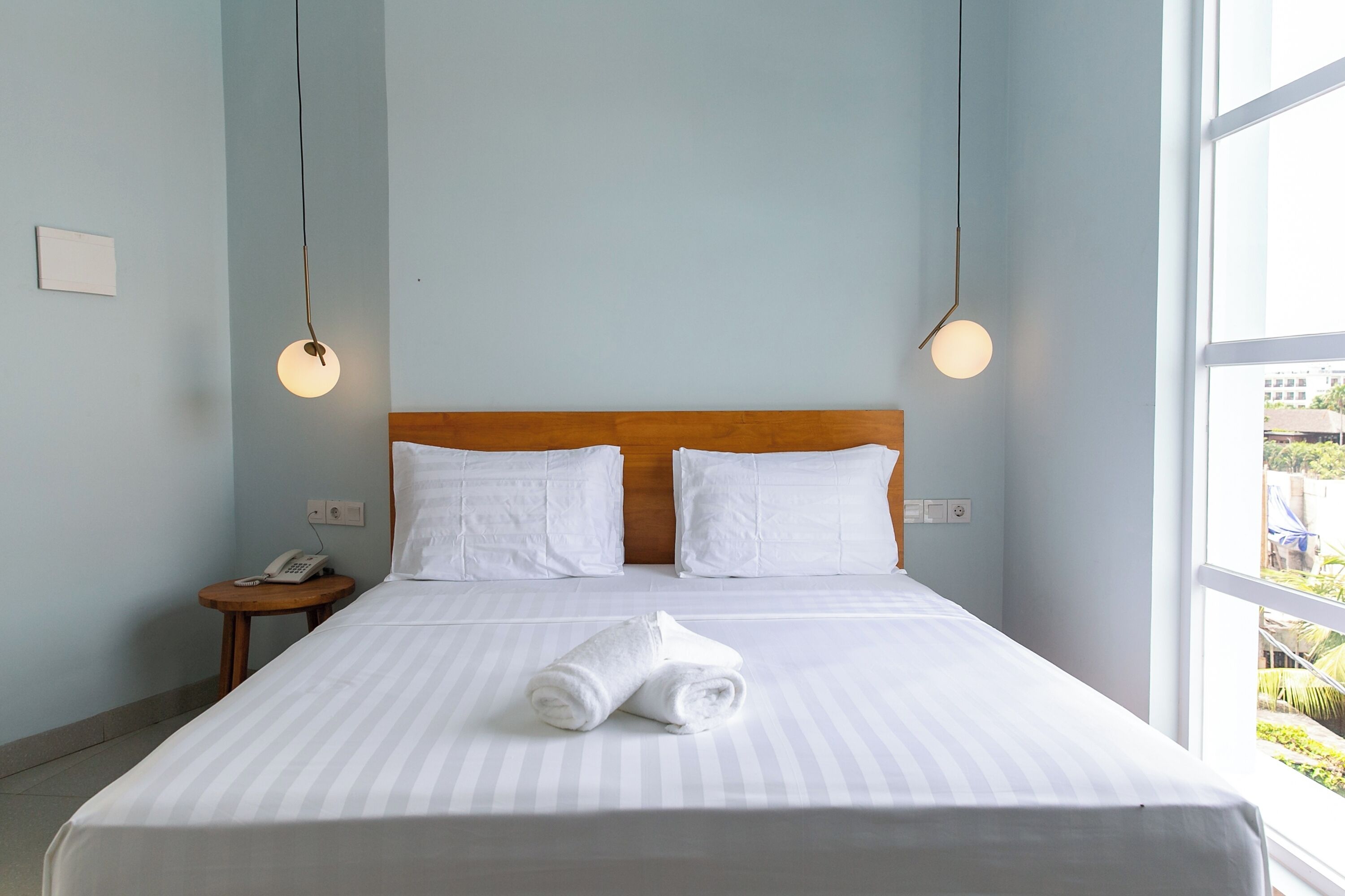 A hotel room with a large bed, two white pillows, a wooden headboard, a bedside table with a phone, and a window with a view of greenery.
