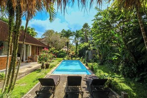 A rectangular swimming pool with a clear blue water surface, surrounded by lush greenery and palm trees. There are two lounge chairs facing the pool, and a small patio area with a table and chairs to the left. - Taman Lituhayu Cottages Ubud (Ubud)