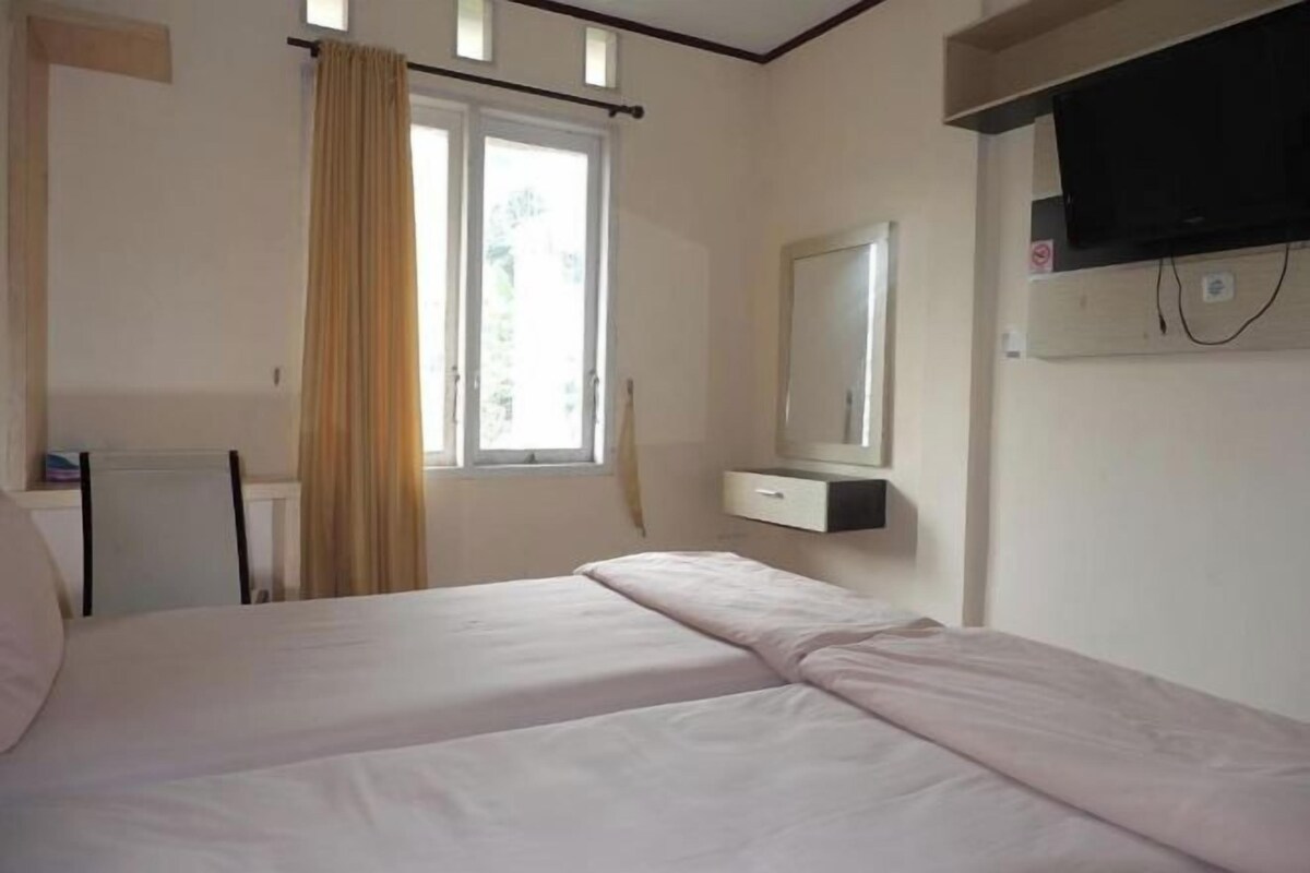 A room with a bed, a desk, a chair, a window with curtains, a television mounted on the wall, and a mirror.