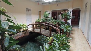 Um jardim coberto e interno com uma ponte de madeira sobre um espelho d'água, cercado por diversas plantas e um piso de azulejos.