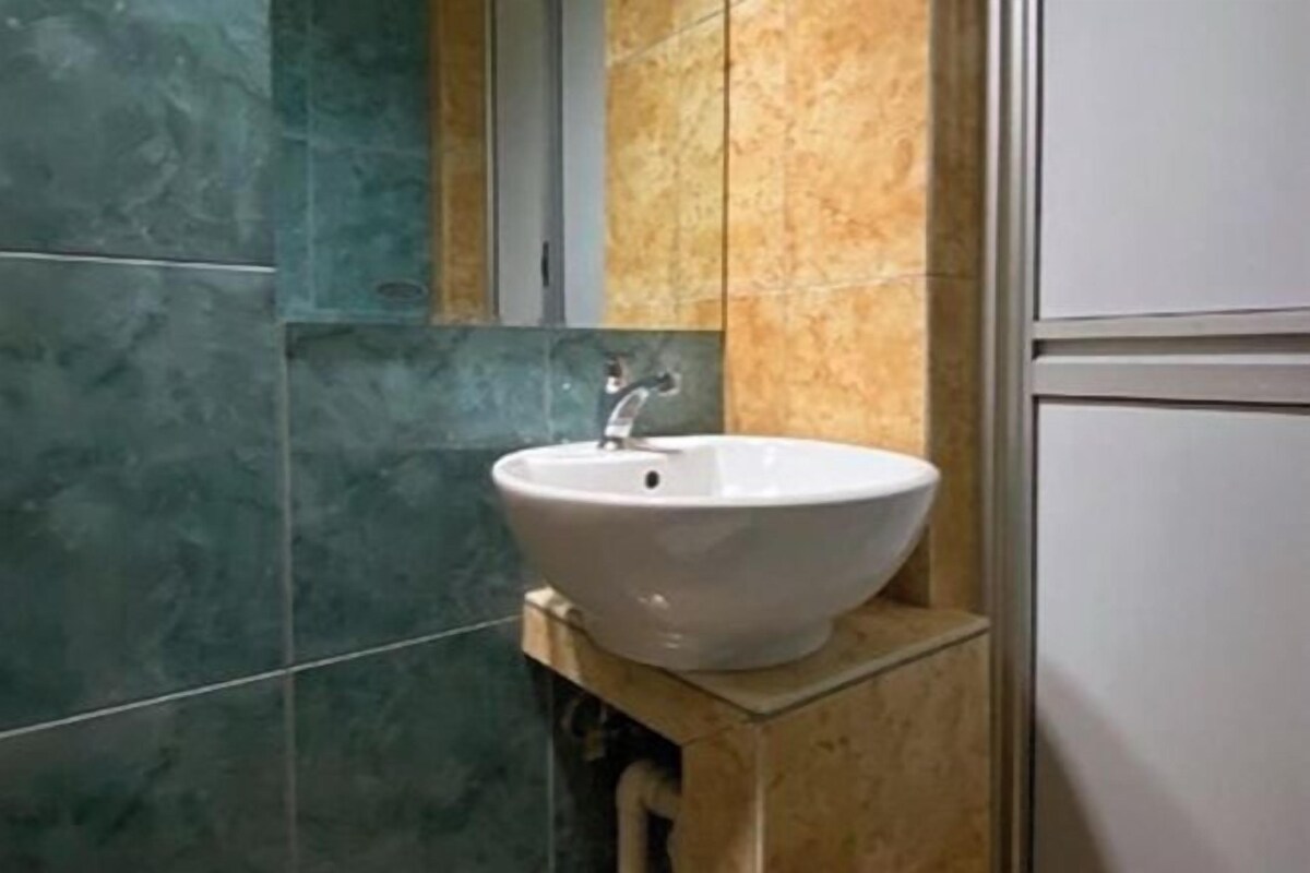 Une salle de bain avec un lavabo blanc posé sur un meuble-vasque en bois, des murs carrelés aux tons de vert et de beige, et un miroir au-dessus du lavabo.