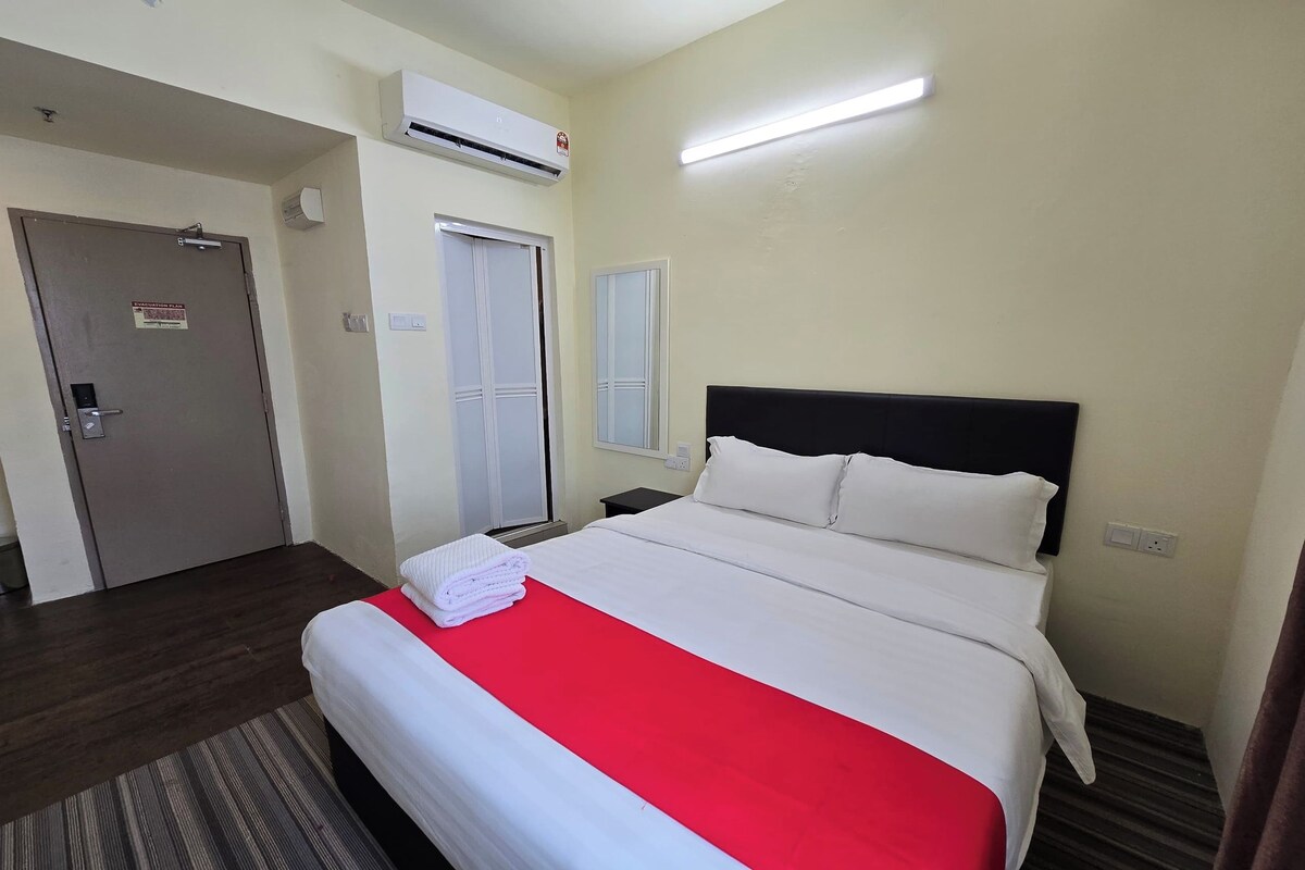 Una habitación de hotel con cama, colcha roja, cabecera blanca y equipo de aire acondicionado en la pared.