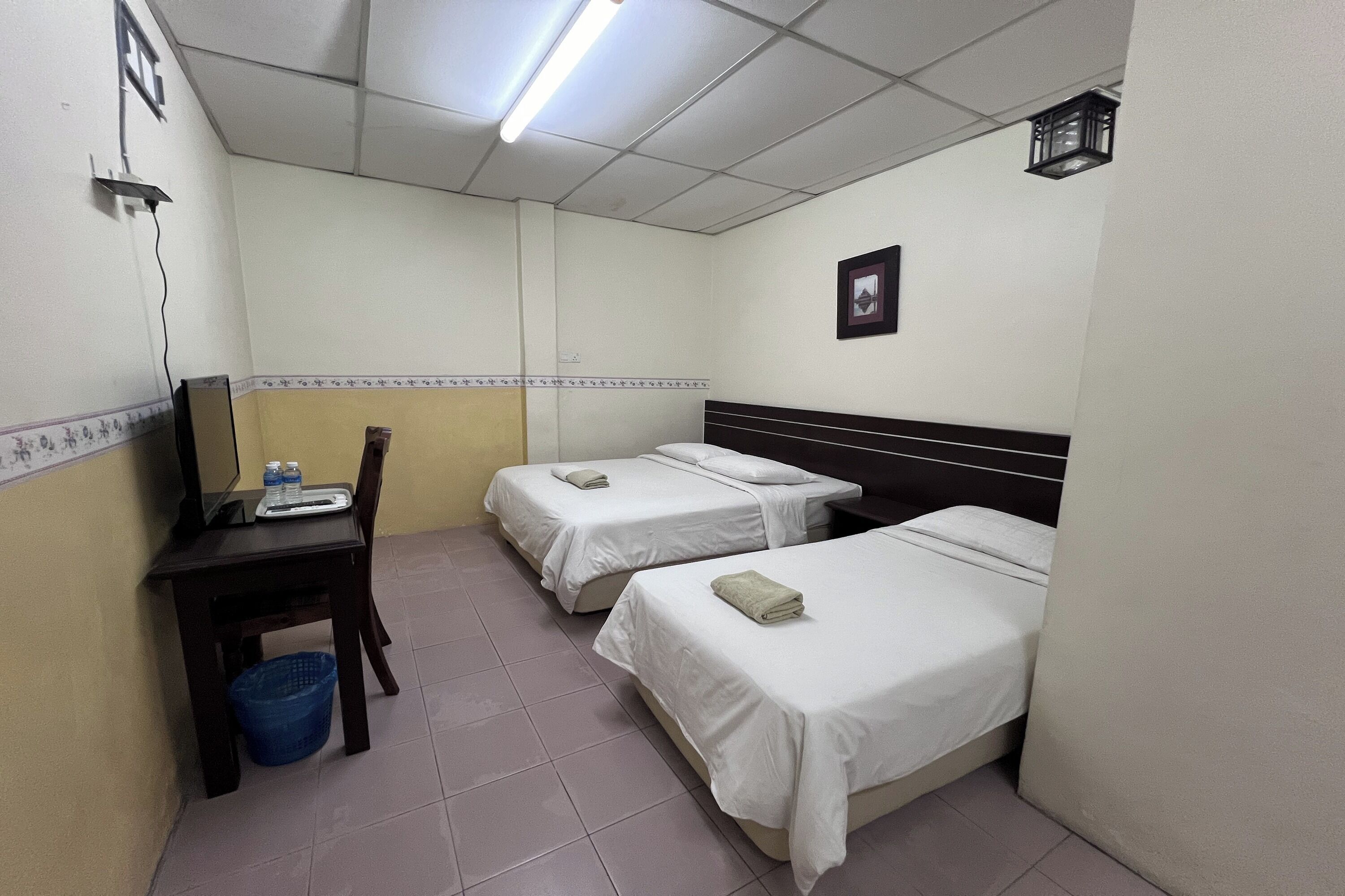 Quarto de hotel com duas camas de solteiro, uma escrivaninha com cadeira, um cesto de lixo azul e uma pequena mesa com garrafas de água.