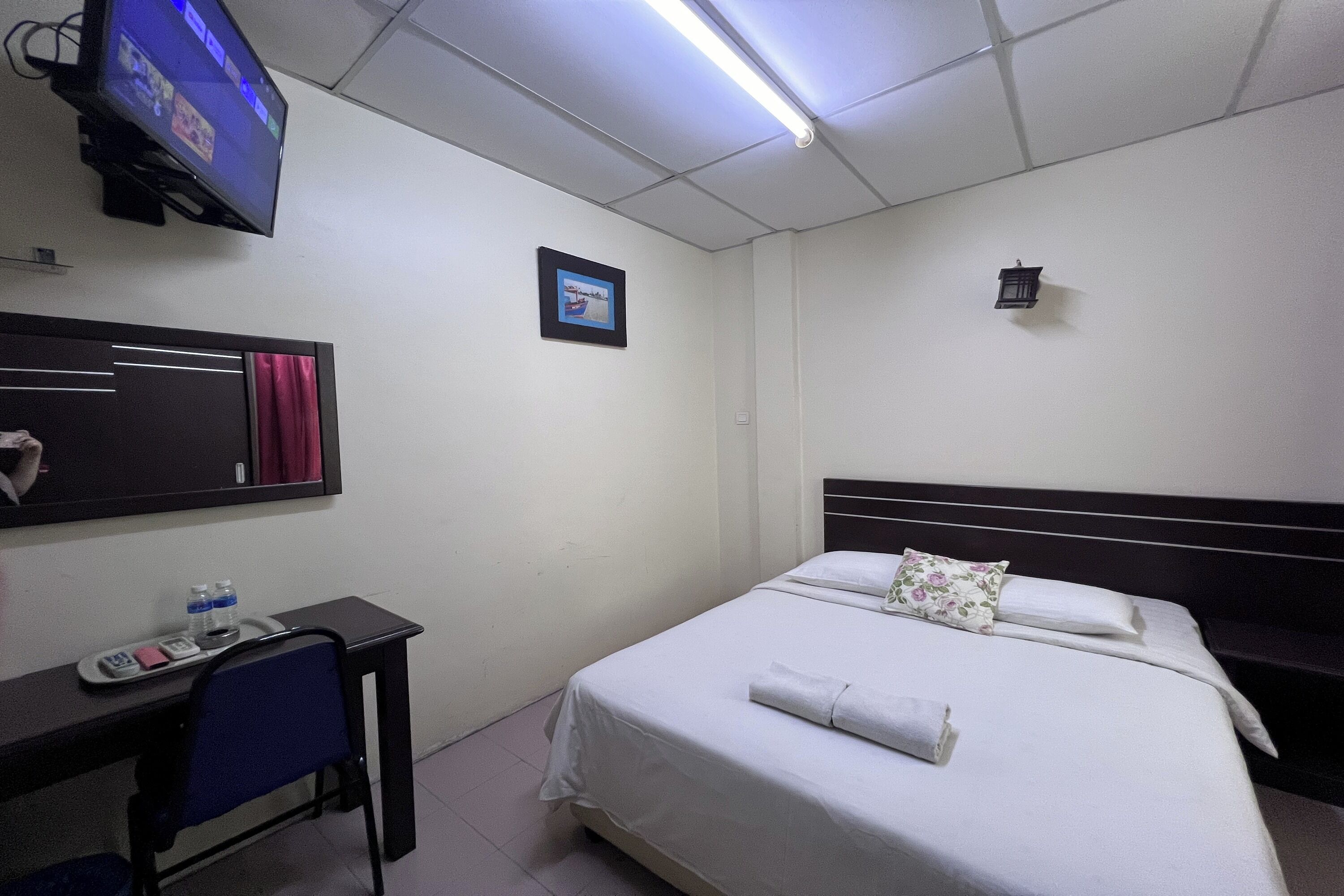 Habitación de hotel con una cama, un escritorio con silla, un televisor montado en la pared y un pequeño marco de fotos.