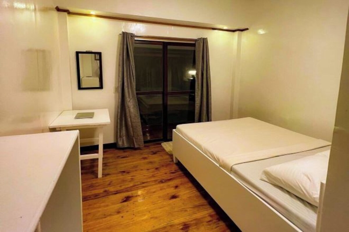 A small, clean room with a wooden floor, a single bed, a desk, and a window with curtains.