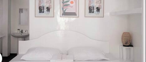 A modern bedroom with a white bed, a bedside table, a wall-mounted sink, and three framed artworks.