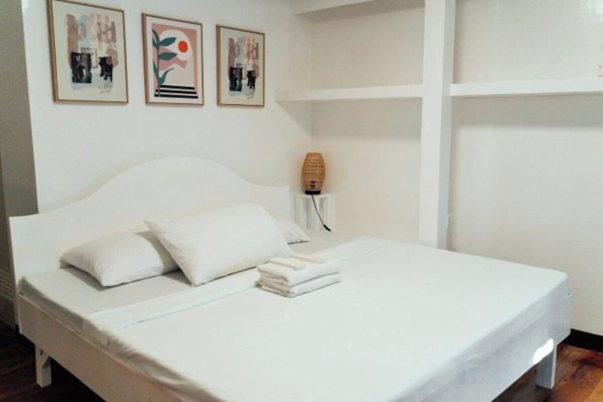 A neatly made bed with white linens and pillows, a bedside lamp, and three framed artworks on the wall.