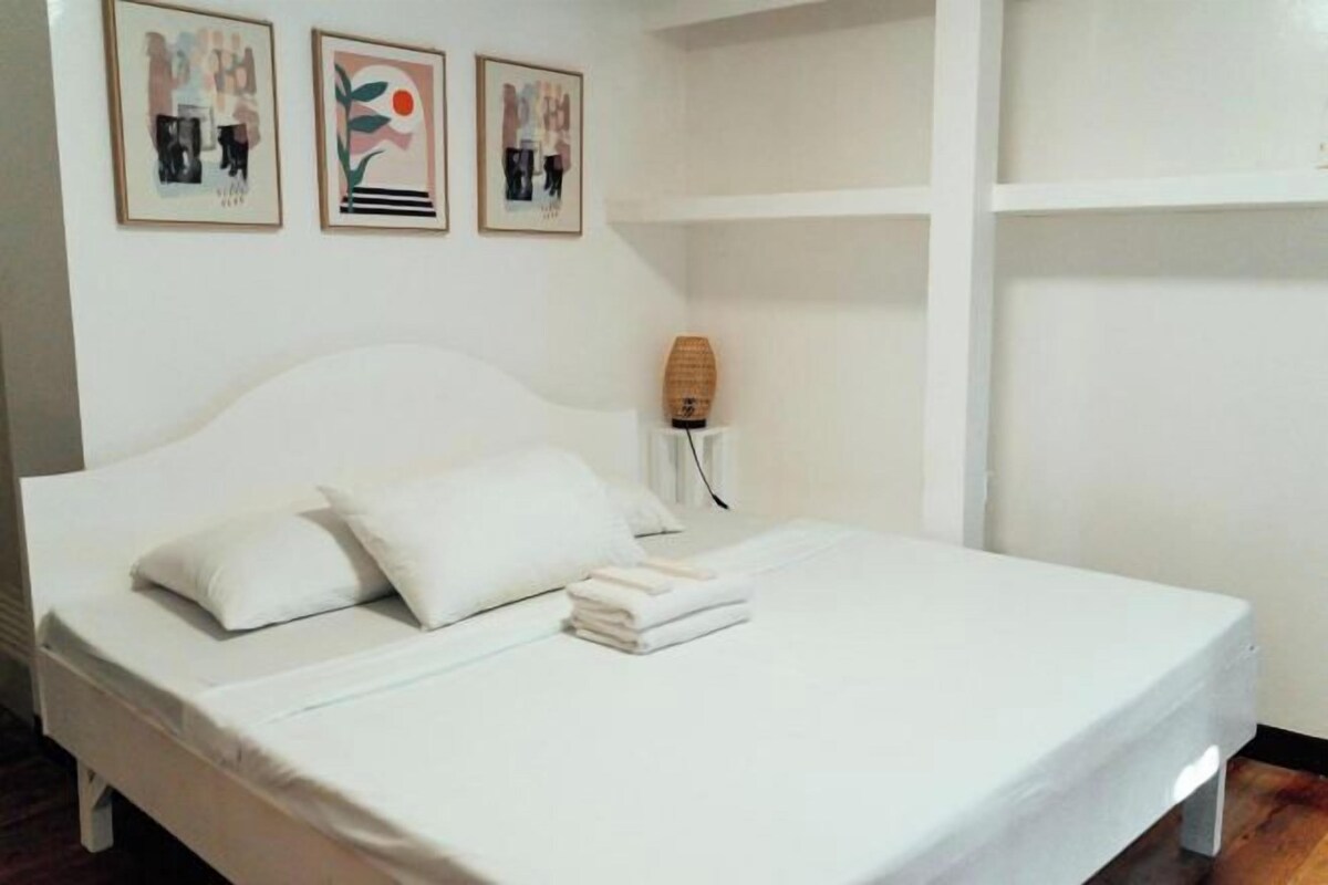 A white bed with two pillows and a folded towel, a bedside lamp, and three framed artworks on the wall.