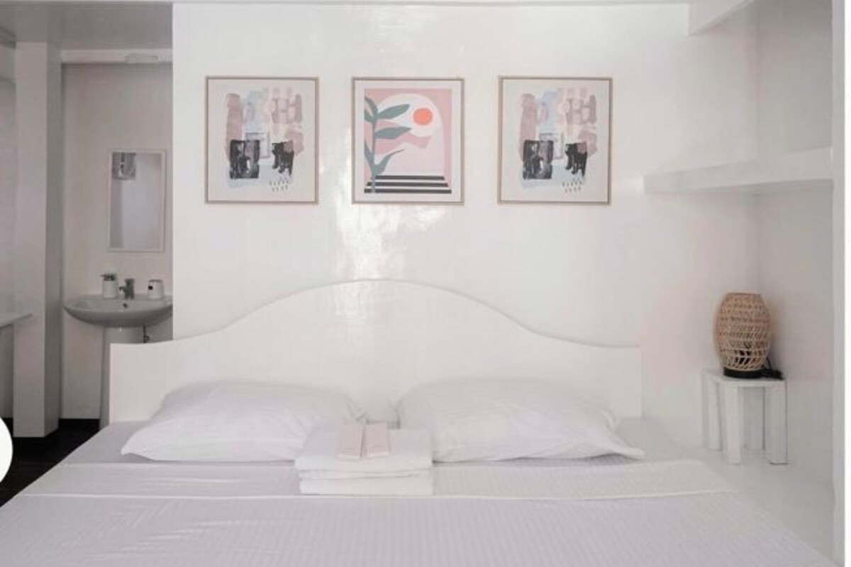 A hotel room with a large bed, a bedside table, a wall-mounted sink, and three framed artworks above the bed.