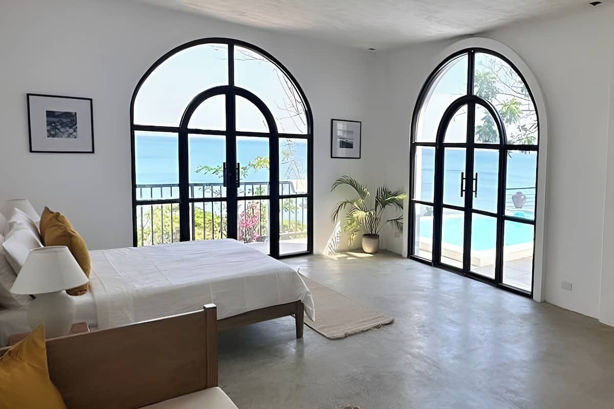 A bedroom with a large bed, two arched windows, a potted plant, and a view of the sea.