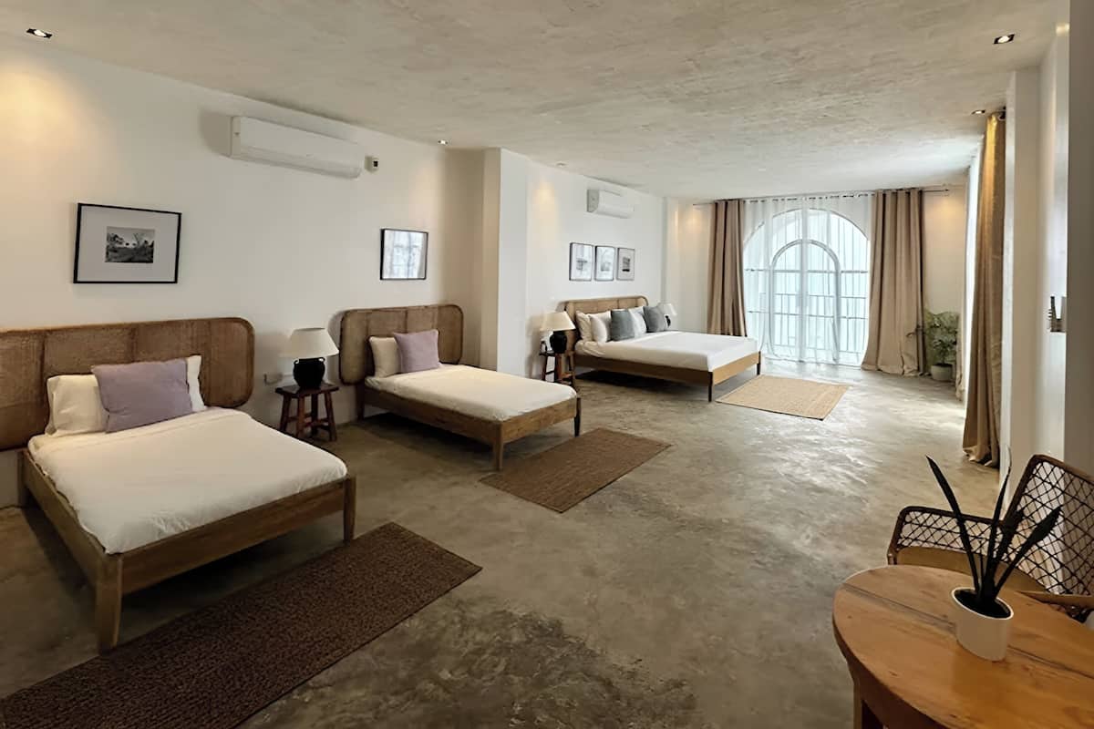 A hotel room with two single beds, a wooden table with a plant, and a large window with curtains.